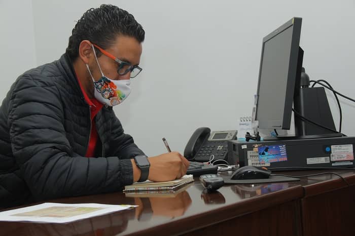 Hombre joven con tapabocas sentado en un puesto de trabajo frente a un computador y escribiendo en una libreta